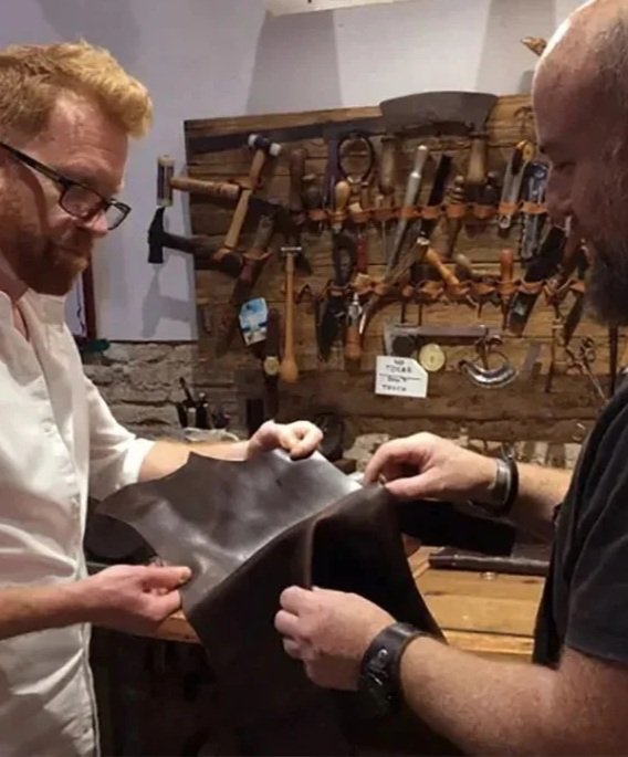 Leather artisan showing a student a piece of leather, with leatherworking tools visible in the background.