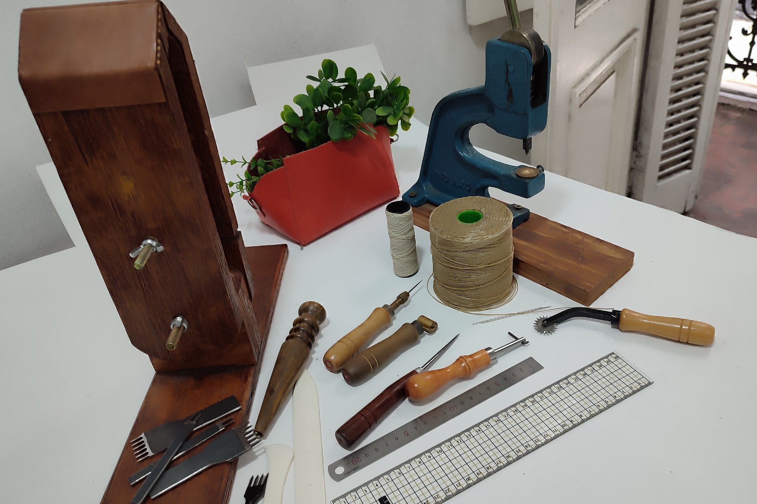 Tools and materials for leatherworking on a white table, including a blue clamp, a wooden ruler, various carving tools, a roll of string, a spool of thread, and a small potted plant in a red container.