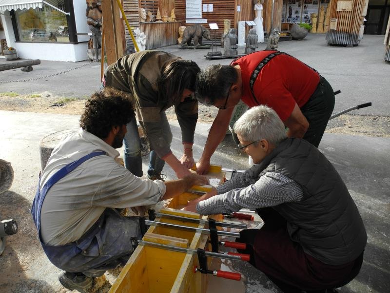 Four people working together on a bronze project outdoors, using clamps and tools on a wooden piece.