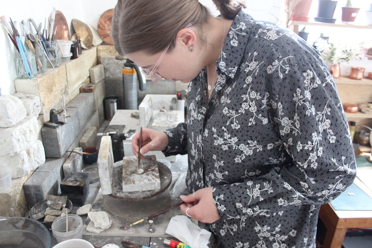 A woman working on a jewelry piece at a workbench in a studio filled with tools and materials.
