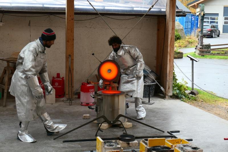 Two people wearing protective suits and masks are working with a high-temperature furnace in a bronzecasting workshop, with one person observing and the other operating the furnace.