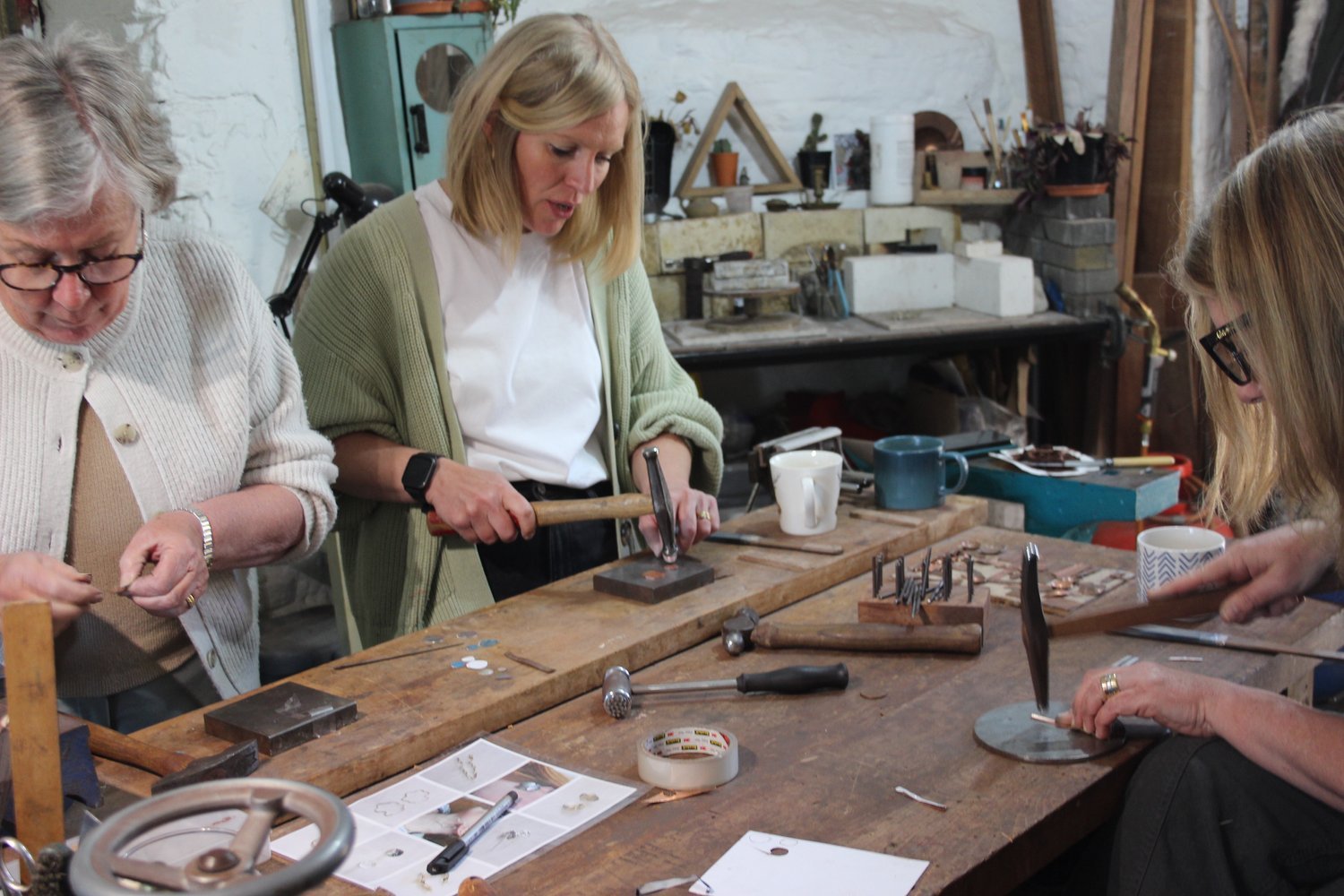 Three women working together in a jewelry workshop, using hammers and tools to create or repair jewelry at a wooden workbench.