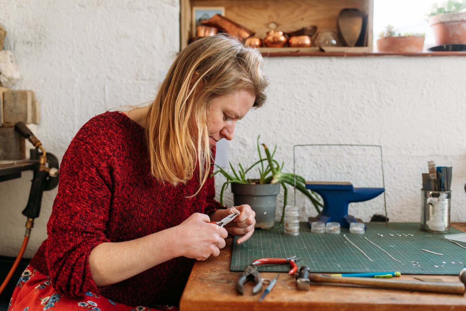A woman with blonde hair wearing a red sweater working on jewelry at a workbench with tools and jewelry supplies.