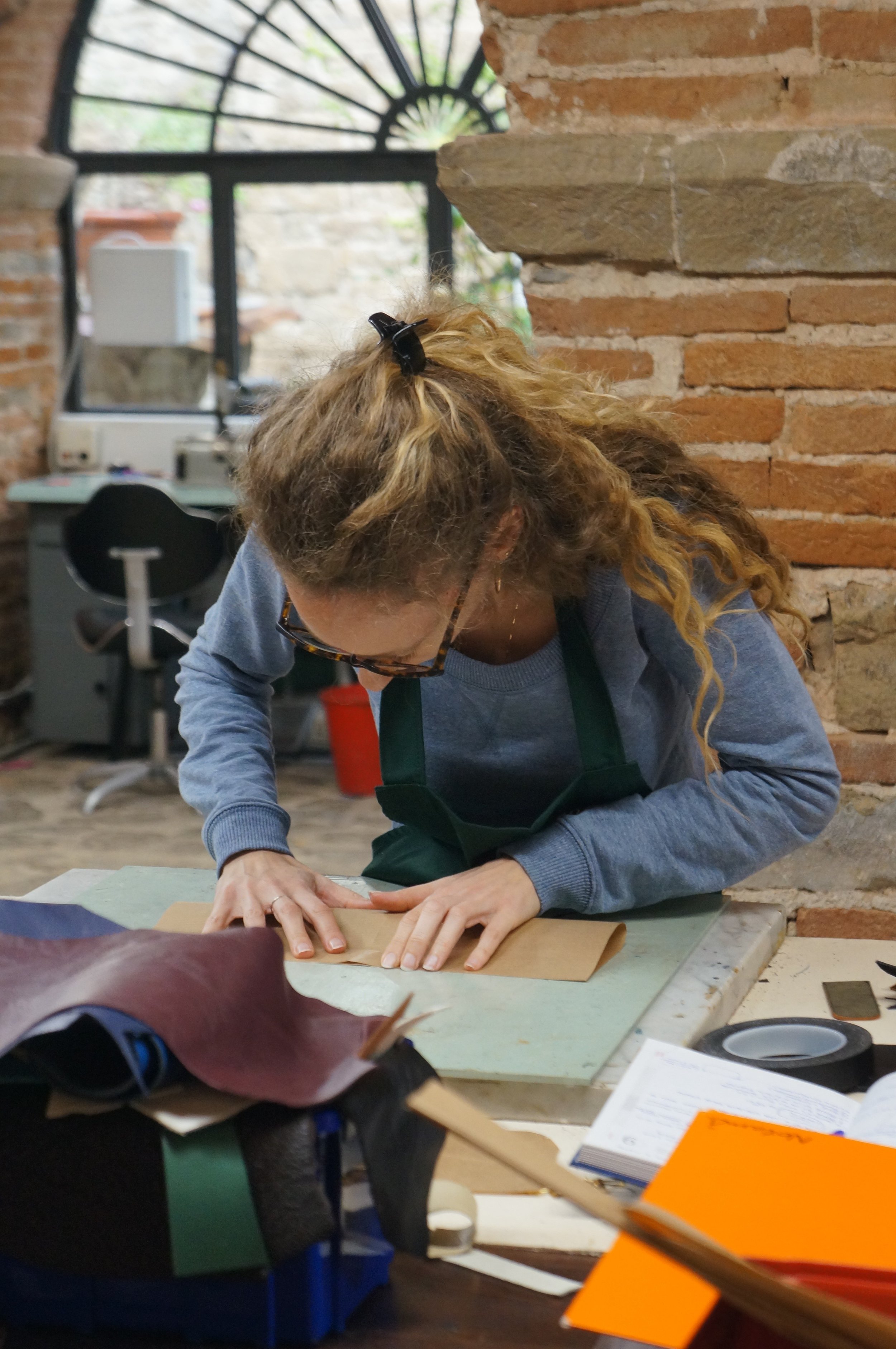 A woman with curly hair tied back, wearing glasses, a gray sweatshirt, and a green apron, bent over a piece of leather on a work table, working on a leather craft project in a workshop with brick walls and a large window.
