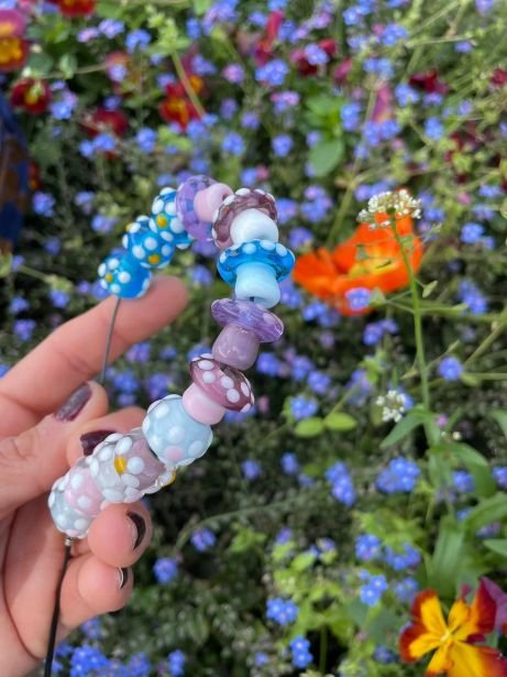 A person's hand with painted nails holding a colorful handmade glass bracelet against a background of purple, blue, yellow, and orange flowers.