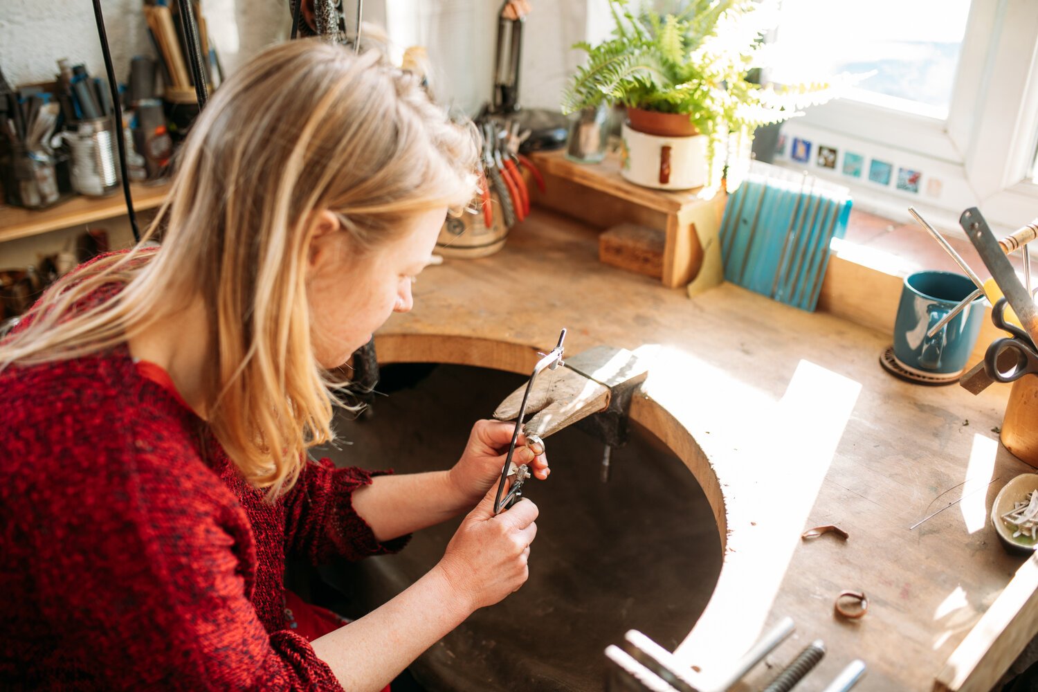 A woman working on silver jewelry making at a woodworking bench with tools and a potted fern in a bright workshop.