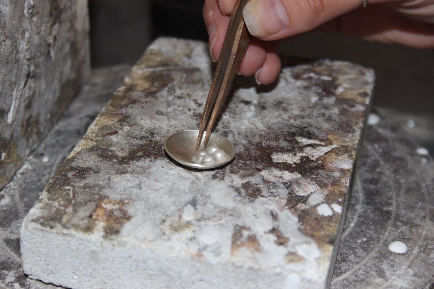A person holding tweezers placing small pearl-like beads onto a metal surface with a dish, on a rough stone workbench in a jewelry workshop