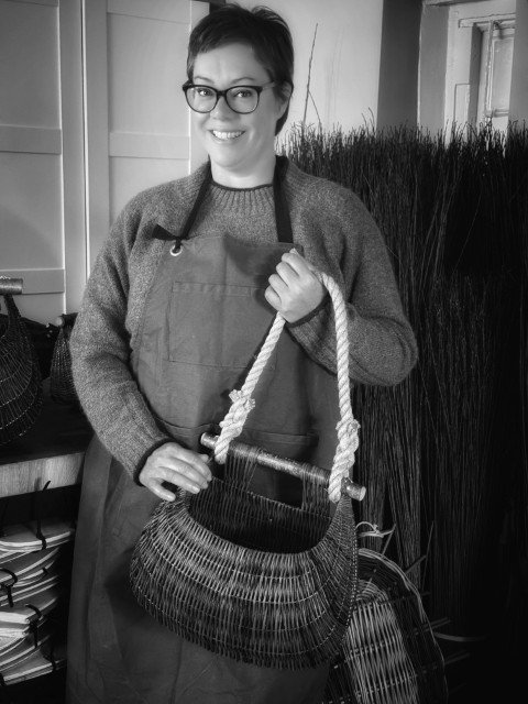 Woman with short hair, glasses, and a smile wearing a sweater and apron, holding a woven basket with a rope handle in a craft shop or workshop.