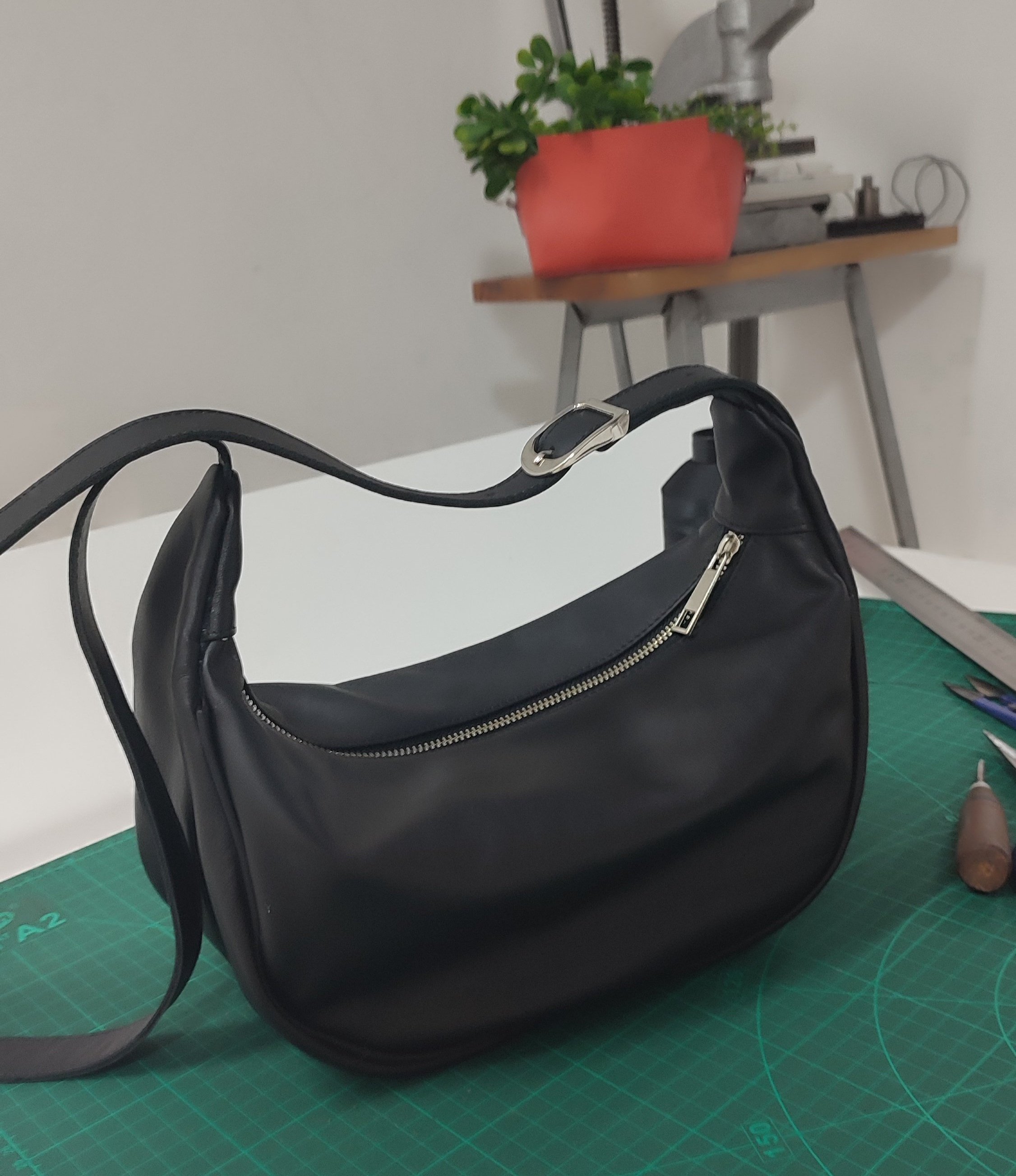 Handmade black leather handbag with silver zipper on a green cutting mat, with tools nearby, in a workspace with a potted plant on a shelf in the background.