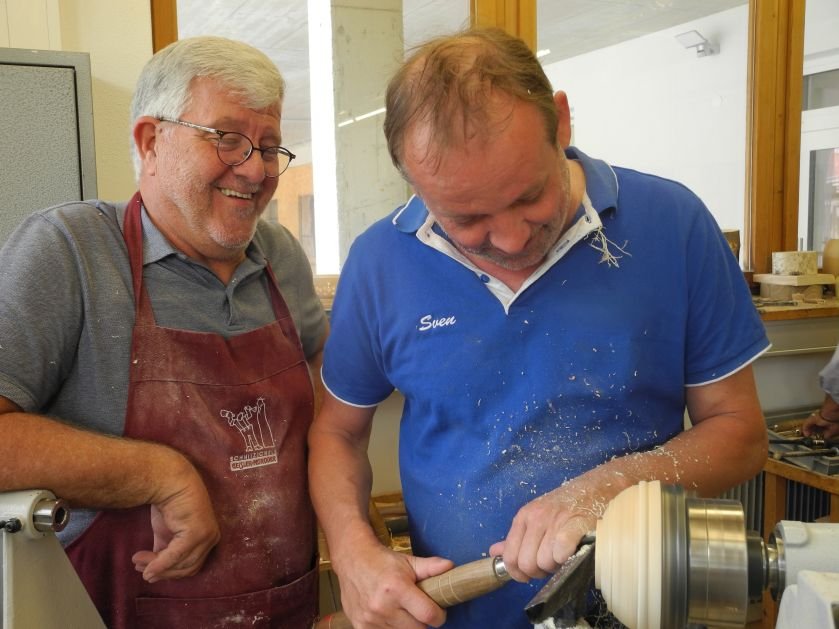 Student shaping wood on a lathe under the guidance of an instructor, both smiling during a woodworking class in Austria.
