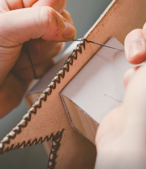 Close-up of hands sewing a piece of tan leather with black thread using a needle.