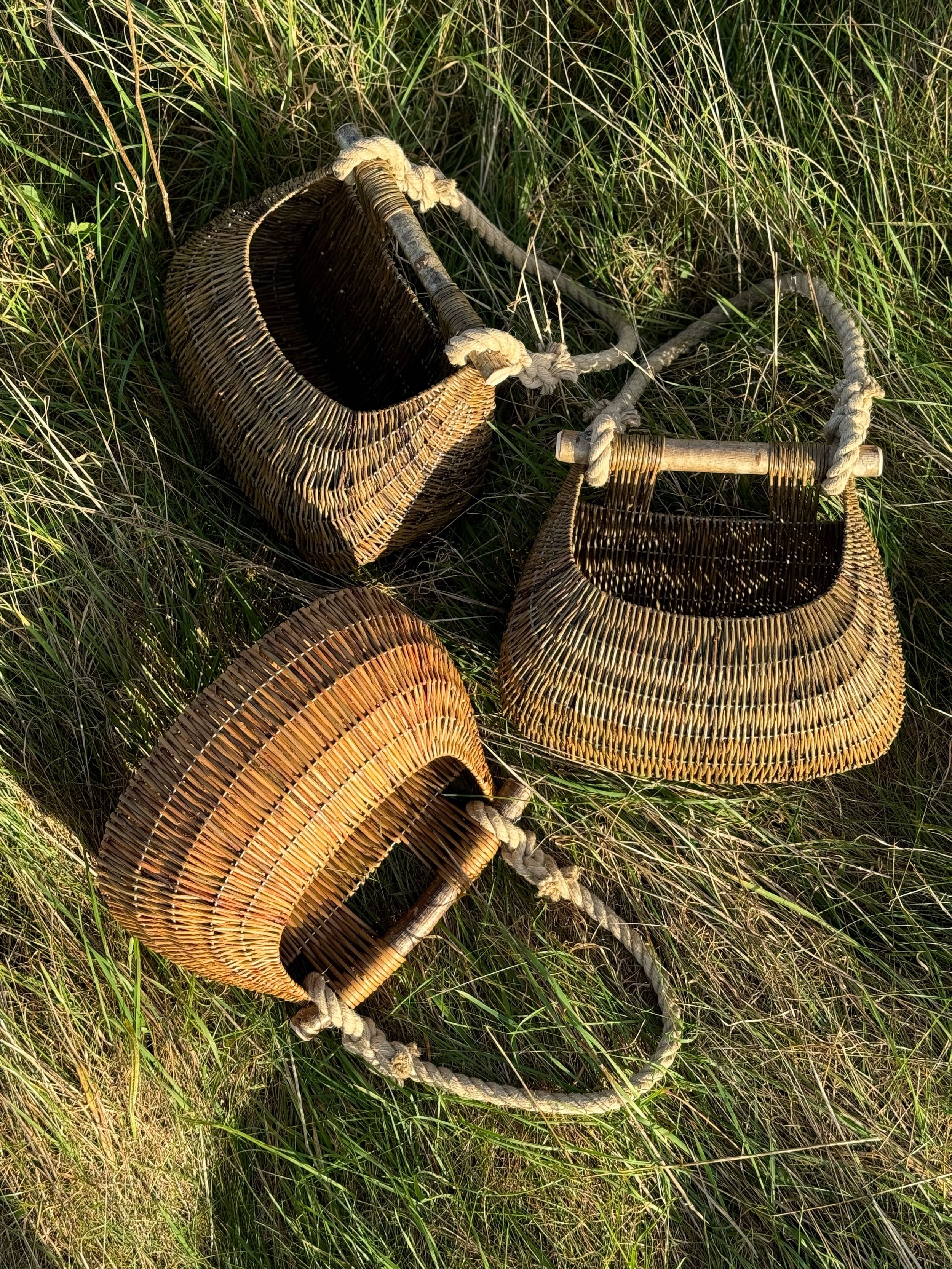 Three handmade woven baskets with wooden handles and knotted ropes, lying on grassy ground.