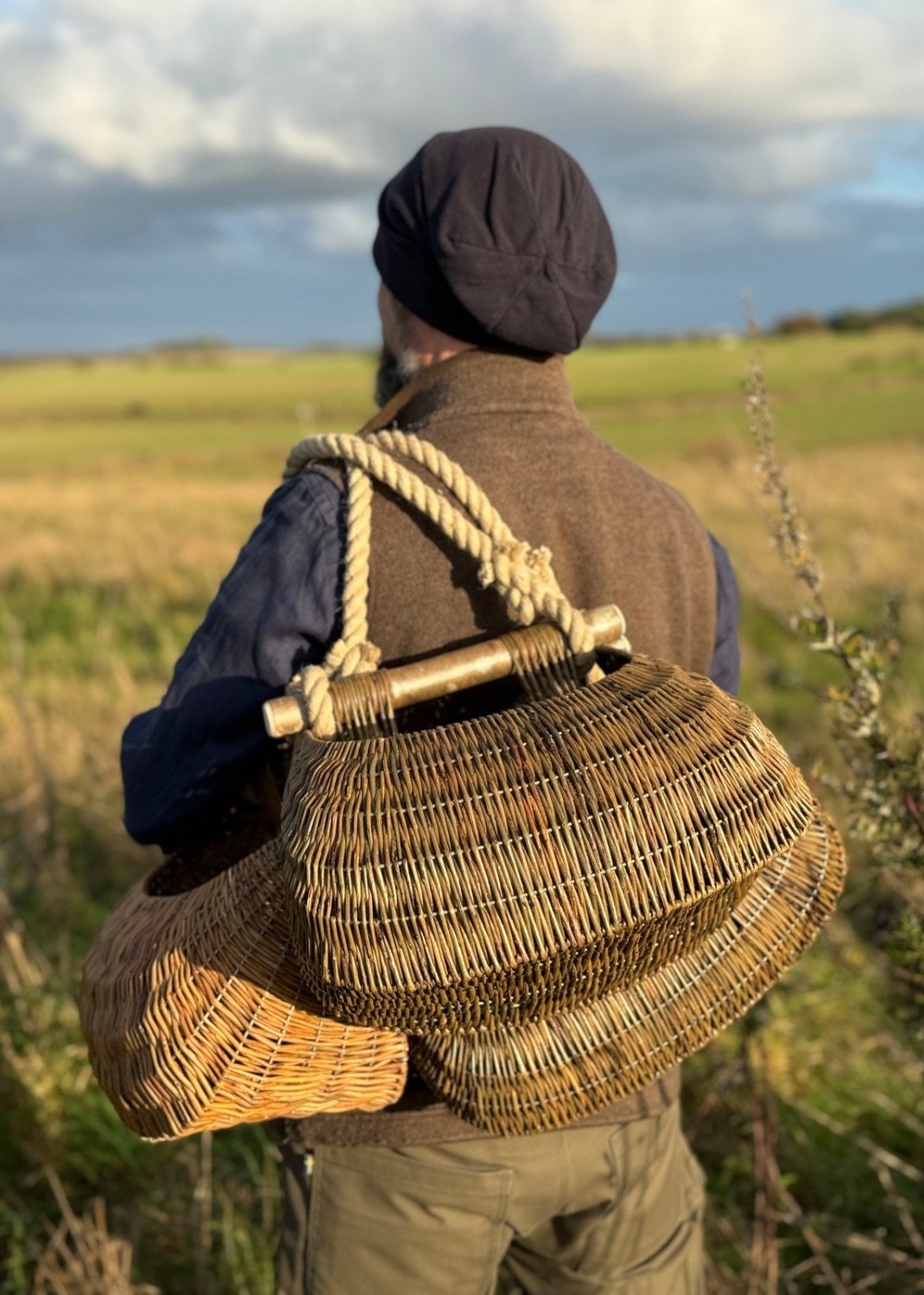 A man wearing a dark hat and vest is standing outdoors in a grassy field, holding three handmade woven wicker baskets on his back with a thick rope. The sky is partly cloudy with some sunlight illuminating the scene.