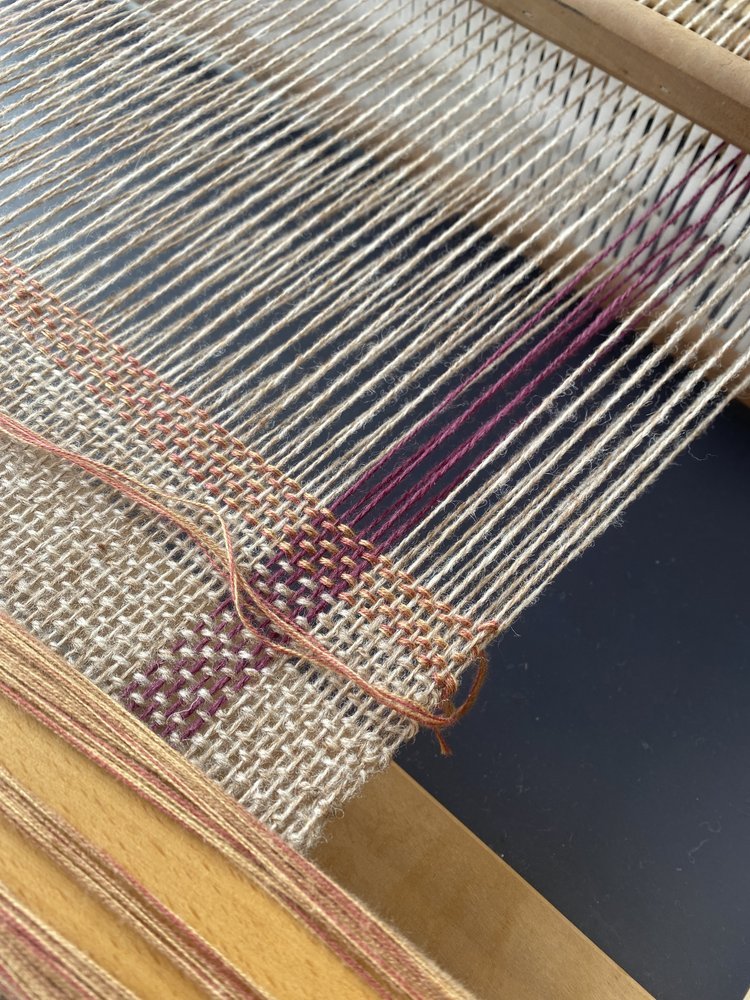 Close-up of a weaving loom with partially woven fabric showing natural and purple threads.