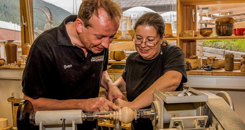 Two women, one man, working on a wood lathe in a woodworking shop filled with wooden bowls and tools, with a window and mountains outside.
