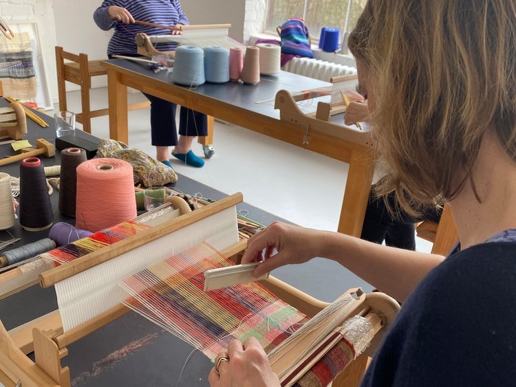 People weaving colorful fabric on a loom in a craft room with various yarns and textiles.