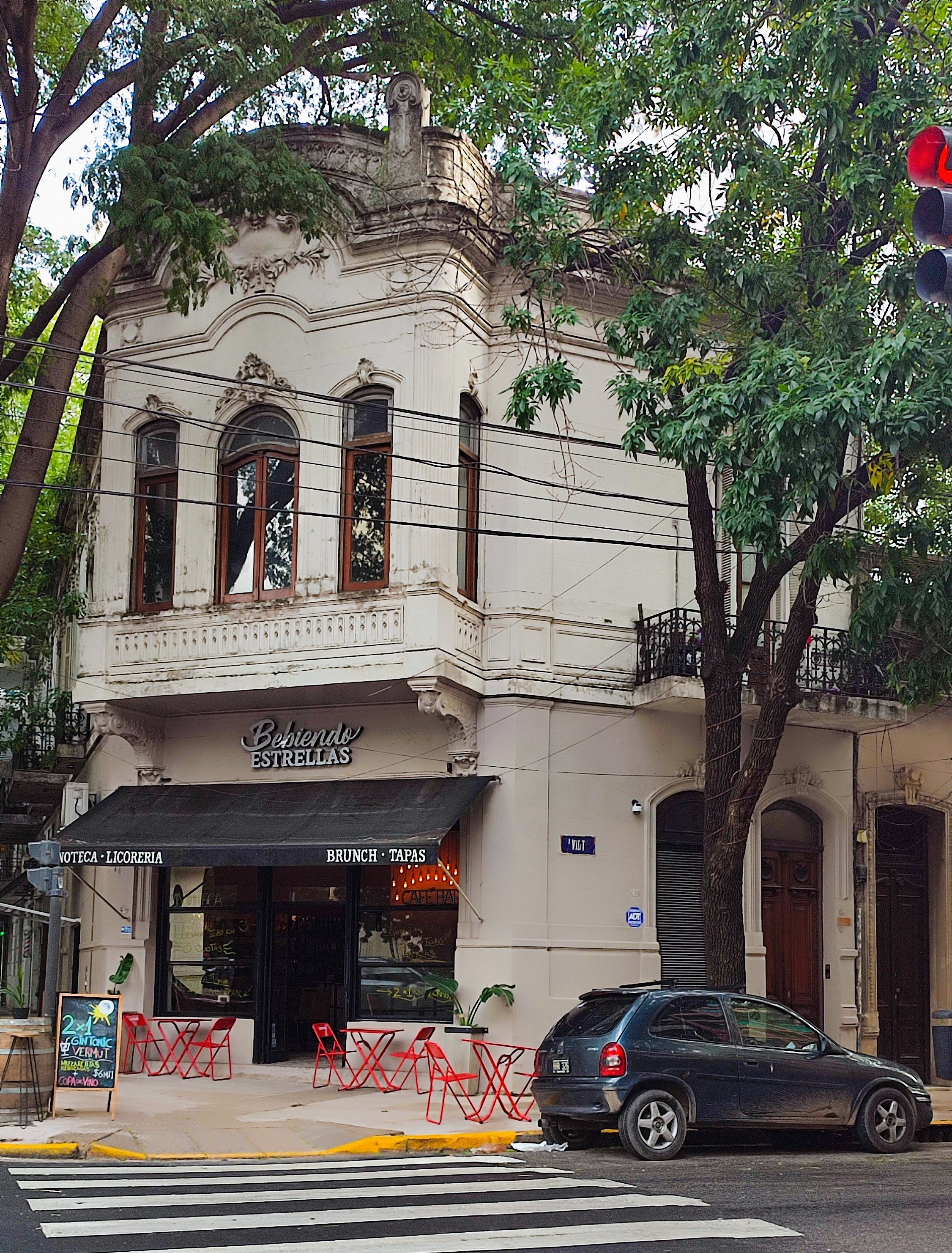 A two-story historic building with ornate architecture houses a café with outdoor red chairs and a chalkboard sign. A car is parked in front, and there are trees and power lines nearby.