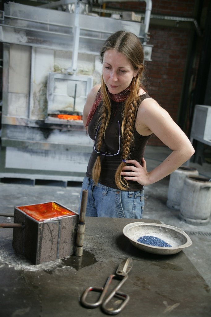 A woman with long braided hair looks at a glassblowing furnace with molten glass inside in a glassblowing studio. There are tools and a bowl of small dark rocks on the work table.