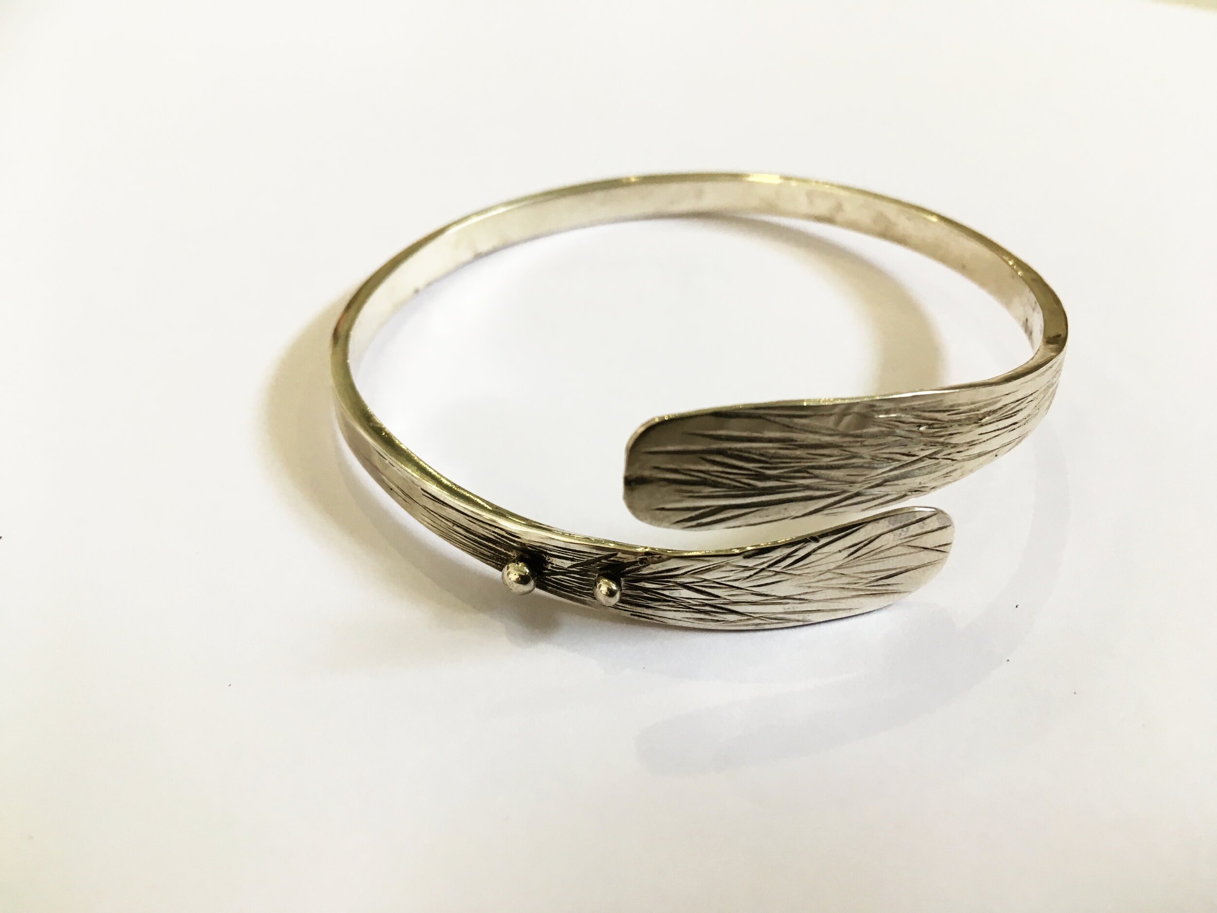 A silver bracelet with textured feather design on a white surface.