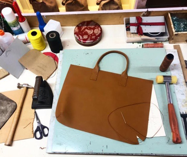 A workspace with a brown leather handbag, sewing tools, thread, scissors, and various crafting supplies.