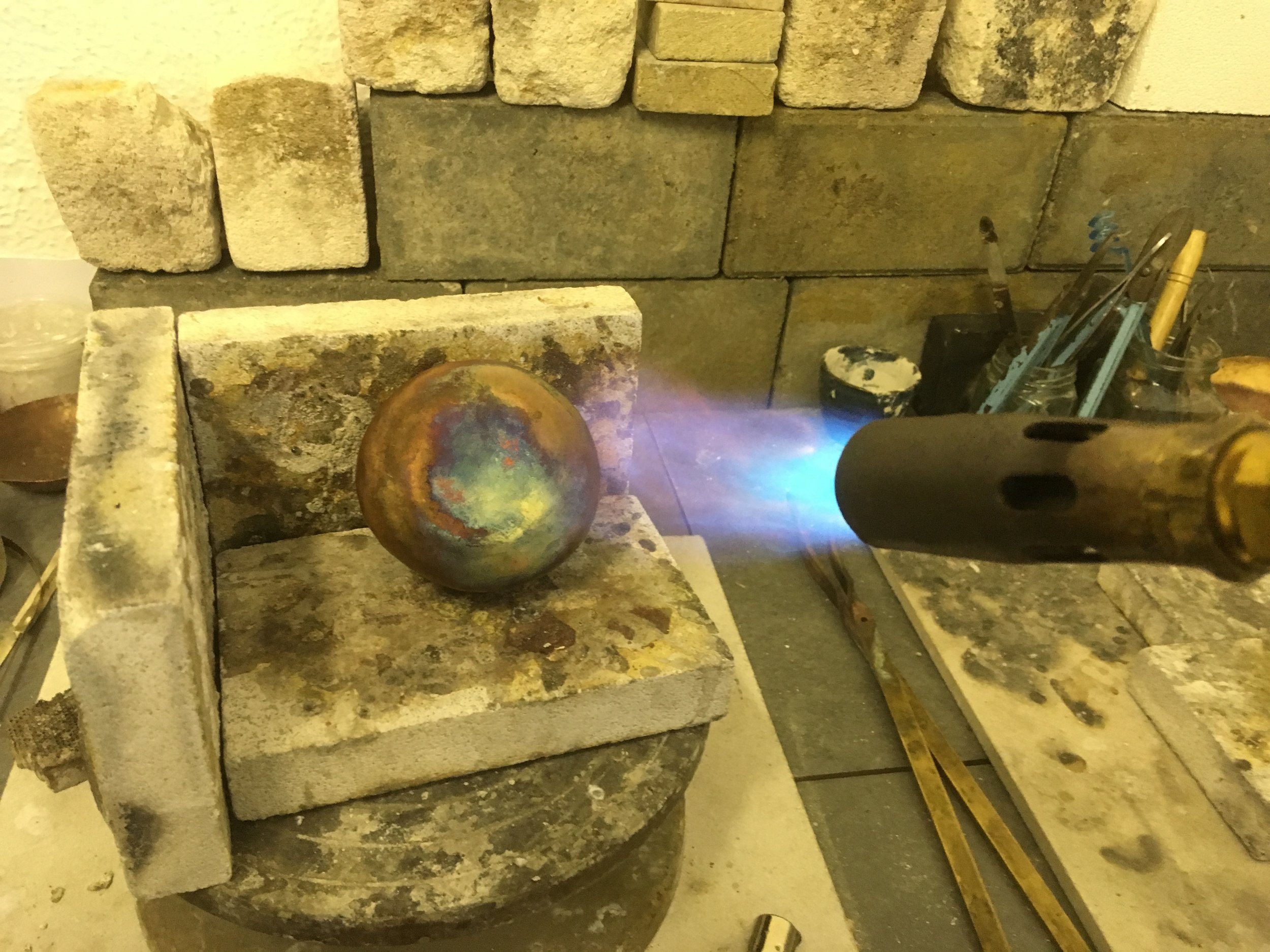 A copper metal bowl is heated with a torch in a metal and brickwork workspace with tools and supplies around.