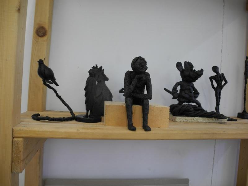 Collection of handmade bronze statues on a wooden shelf, including a bird on a branch, a horse-head figure, a seated figure with a backpack, a mouse-like creature, and a standing abstract figure.