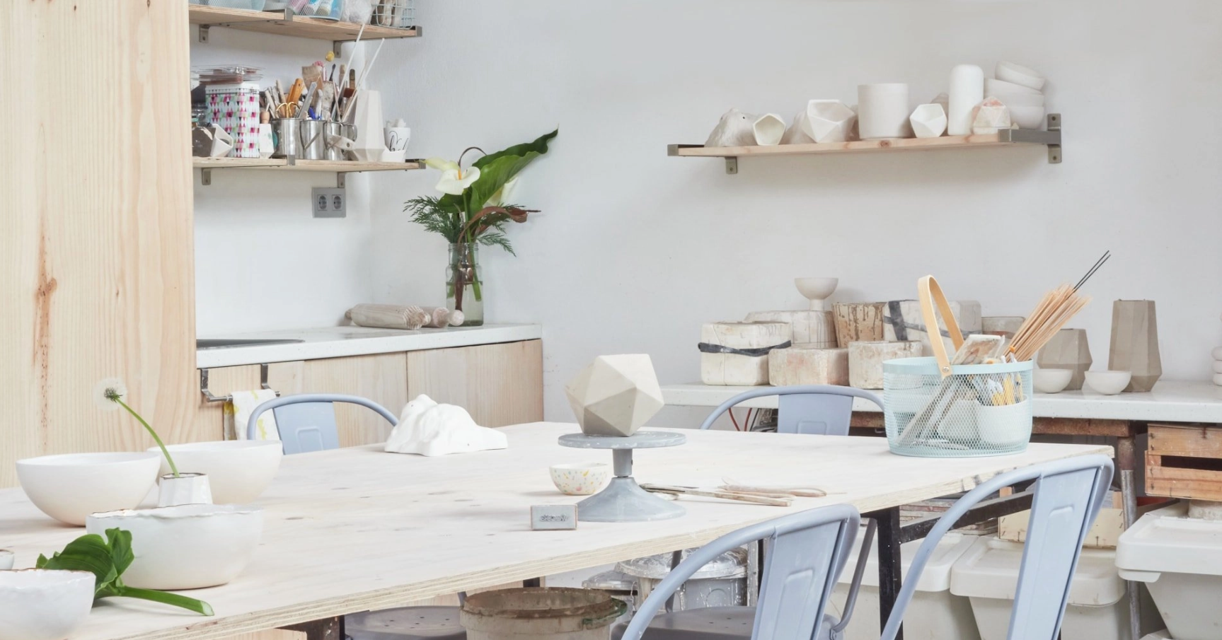 Pottery studio with a worktable, bowls, brushes, and shelves filled with ceramic pieces and supplies.