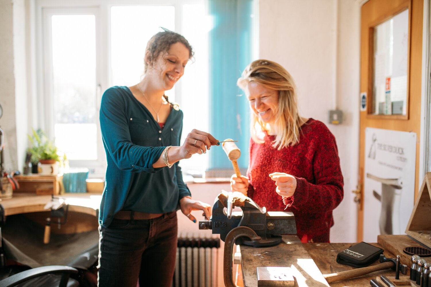 Two smiling women work with jewelry tools in a bright workshop.