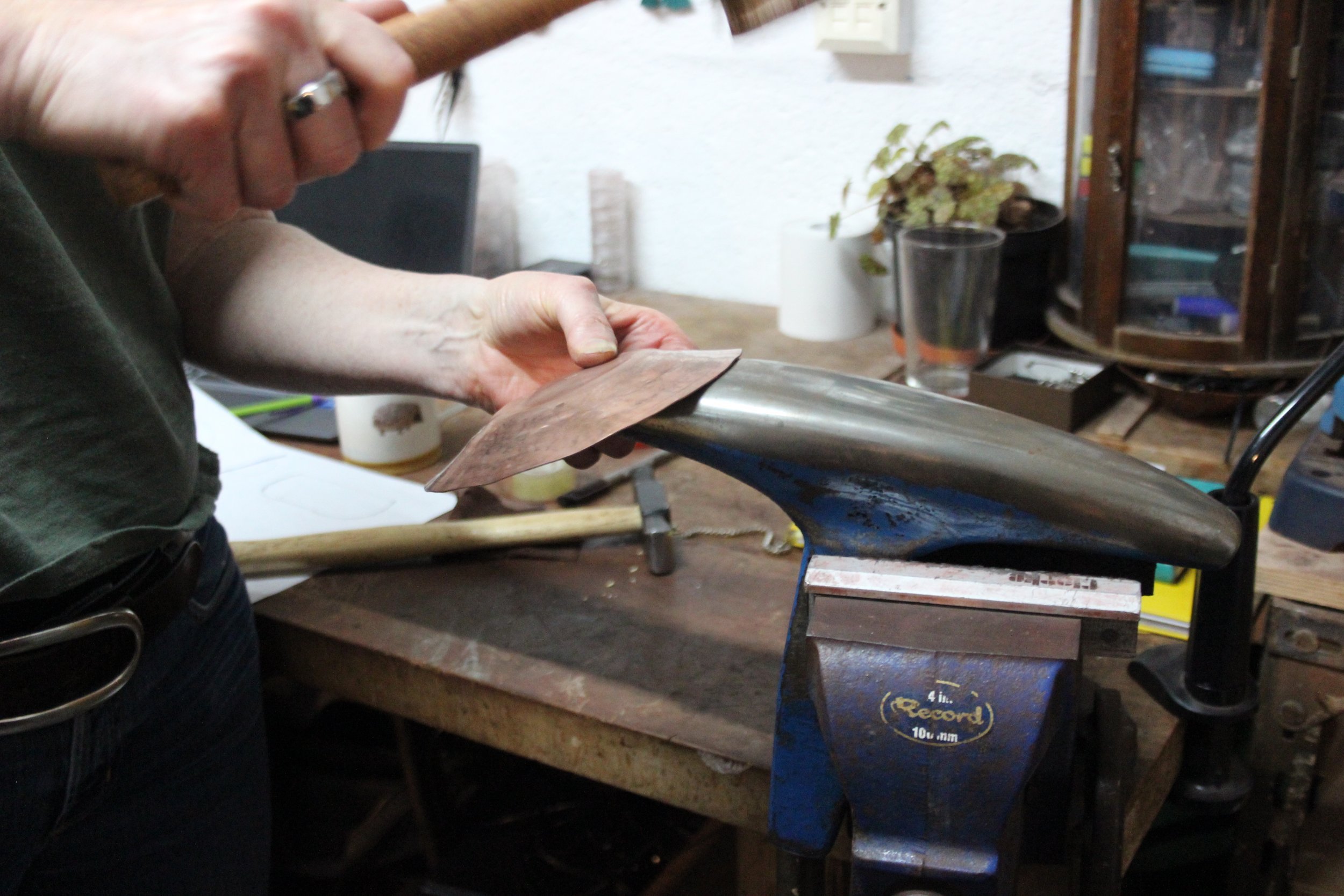 A person hammering a copper bowl into shape on a workbench in a workshop.