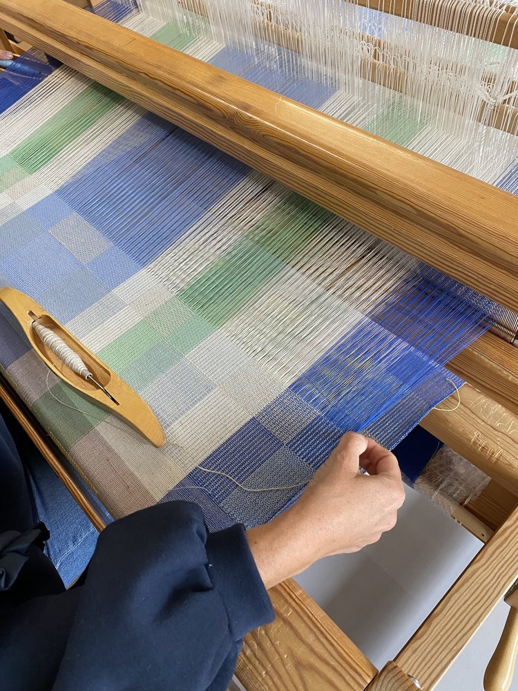 A person working on a loom threading colorful textile threads, with blue, green, and beige hues, creating a patterned fabric.