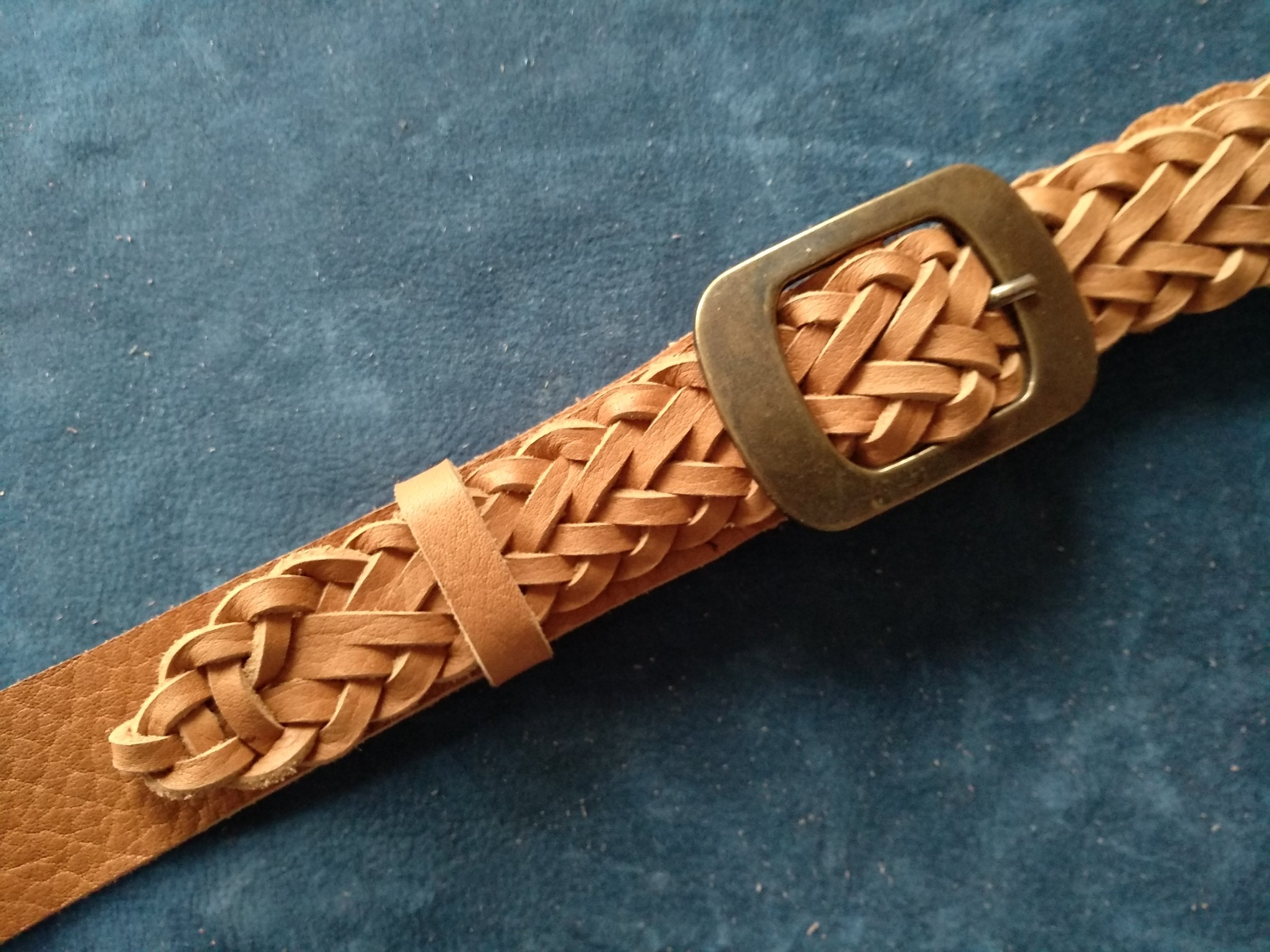 Close-up of a handmade woven tan leather belt with a metal buckle on a blue fabric background.