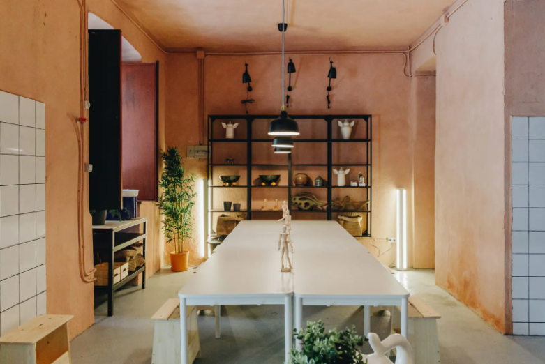Ceramic studio with a long white table, black pendant lights, a black shelving unit with handmade decorative items, and warm ambient lighting in a room with peach-colored walls.