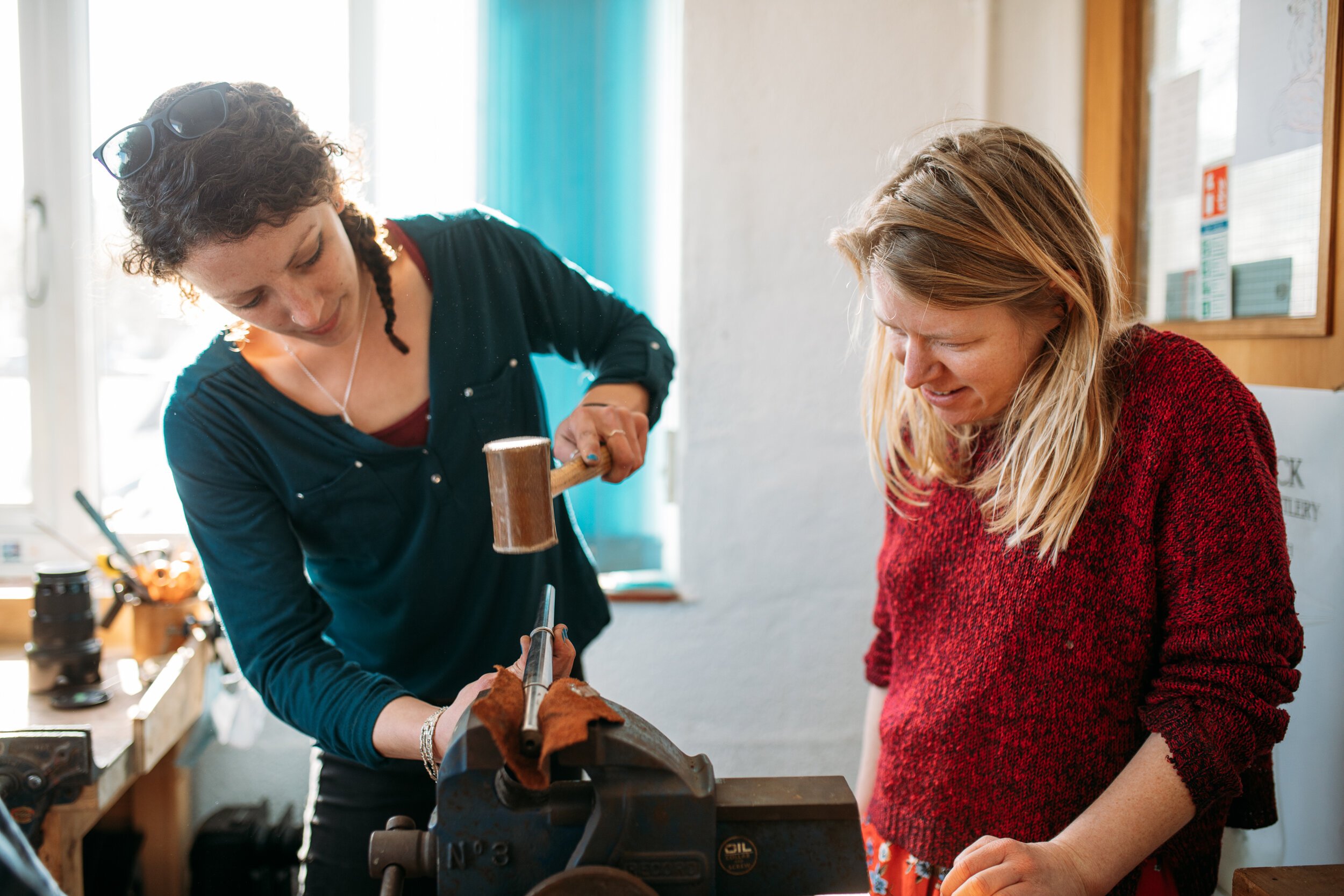 A woman is hammering a piece of wood while another woman watches, inside a workshop with tools and supplies around.