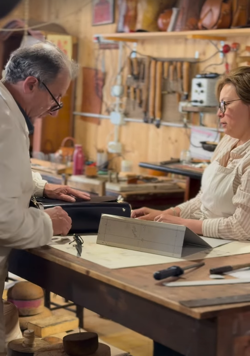A man and woman working in a workshop, handling leather items, with tools and woodwork supplies on the workbench.