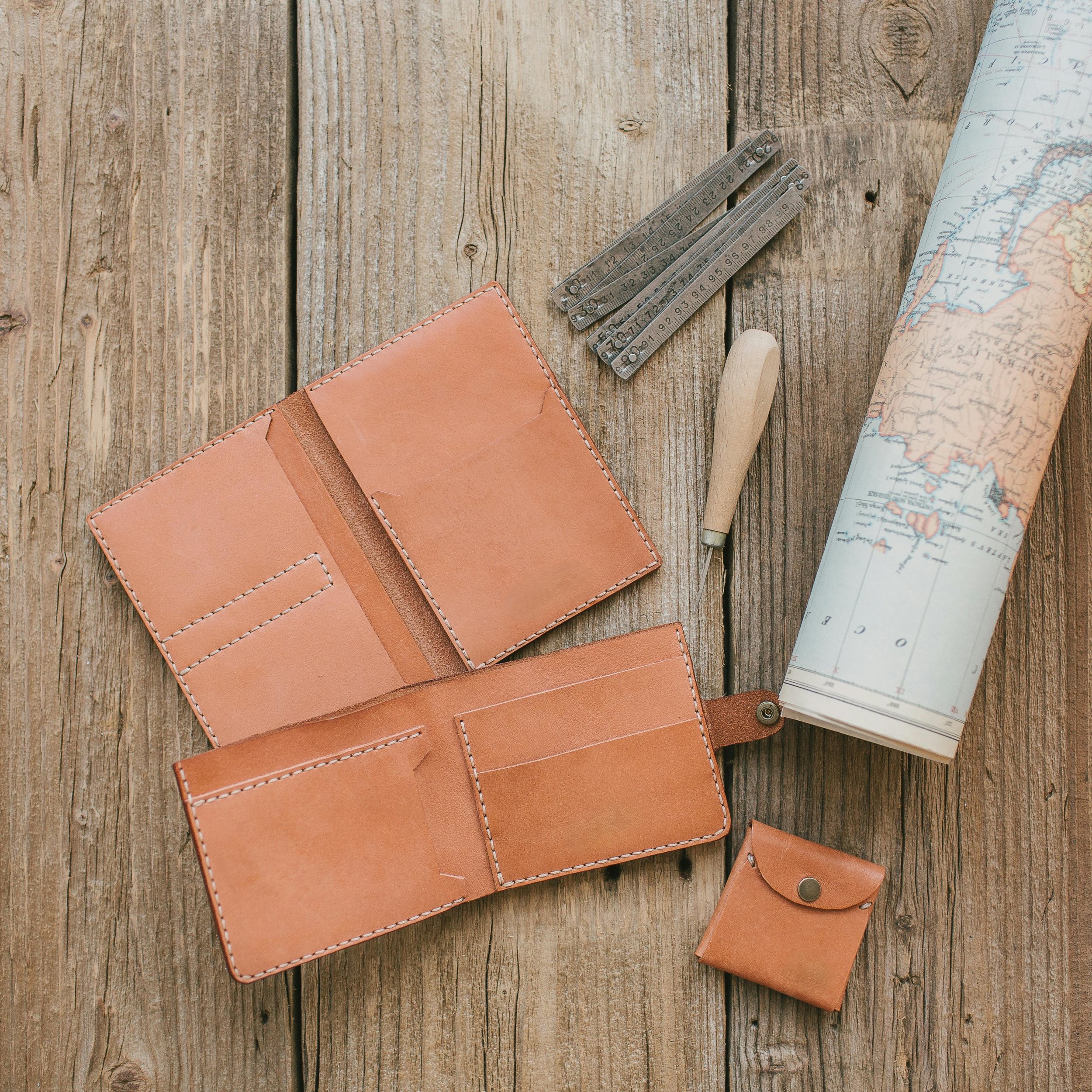 Leather travel wallet, a small leather coin purse, metal measuring tools, a wooden-handled tool, and a rolled-up map on a wooden surface.