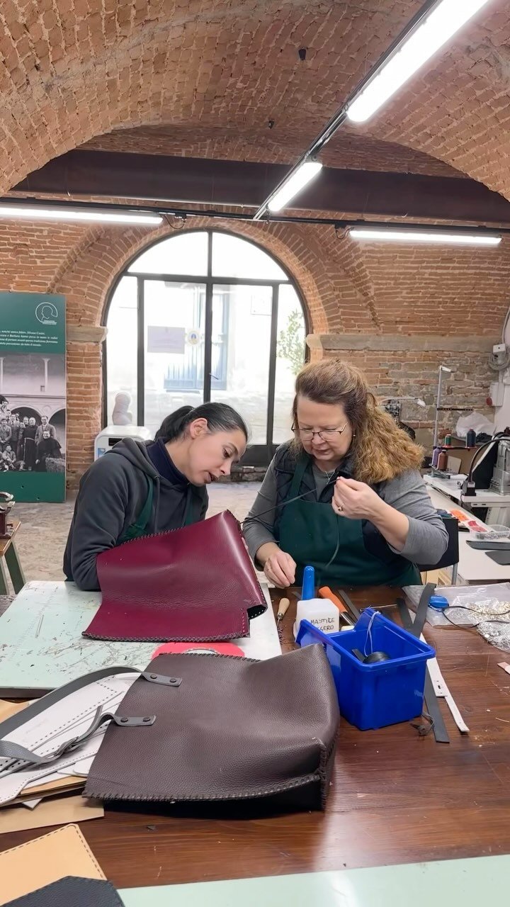 Two women working on leather crafts at a table in a brick-walled workshop, with various tools and bags on the table.