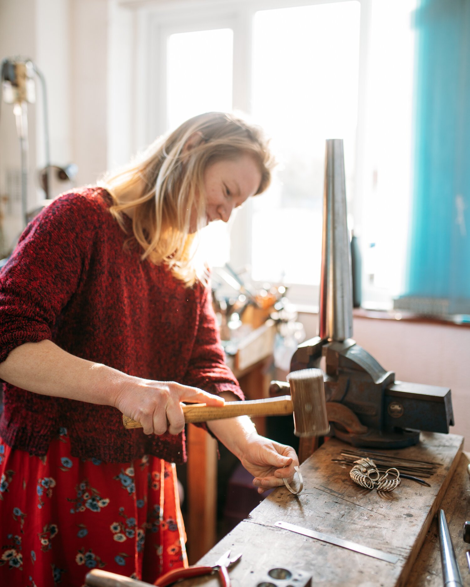 A woman wearing a red sweater and floral skirt is working with jewelry tools at a wooden workbench, using a hammer on a piece of jewelry in a brightly lit studio or workshop.