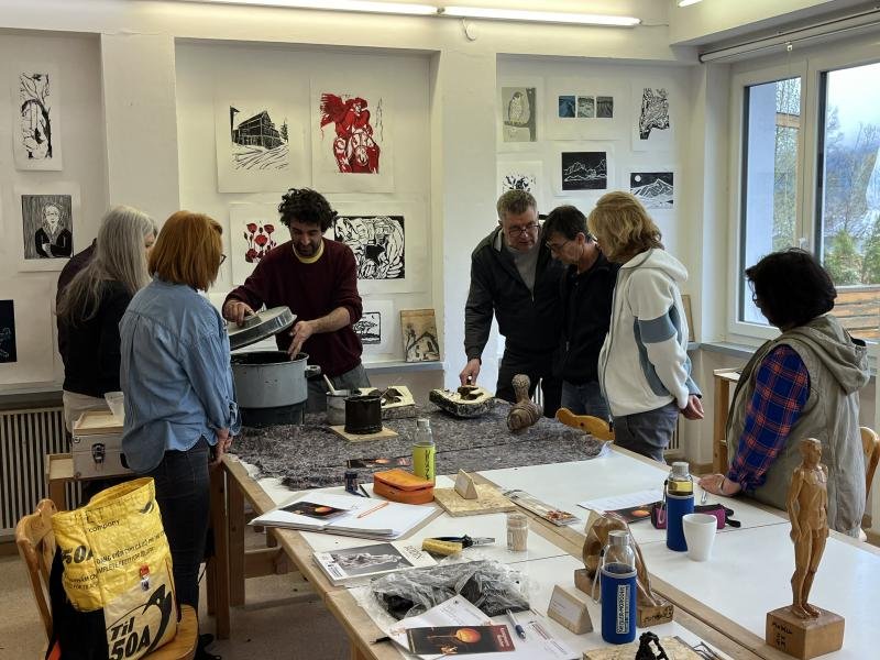 A group of seven people gathered around a table in a bronze casting studio, observing and working on sculptures. The studio has art prints hanging on the walls and a large window letting in natural light.