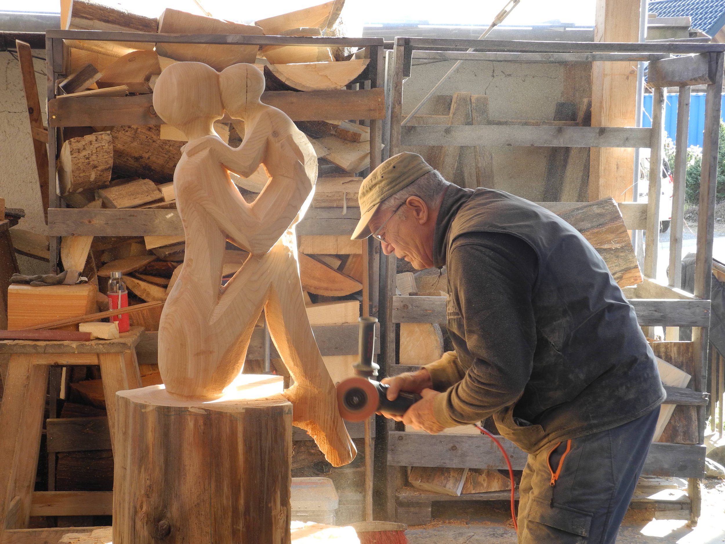 A man working on a wooden sculpture of a couple kissing in a woodworking shop, with wood logs in the background.