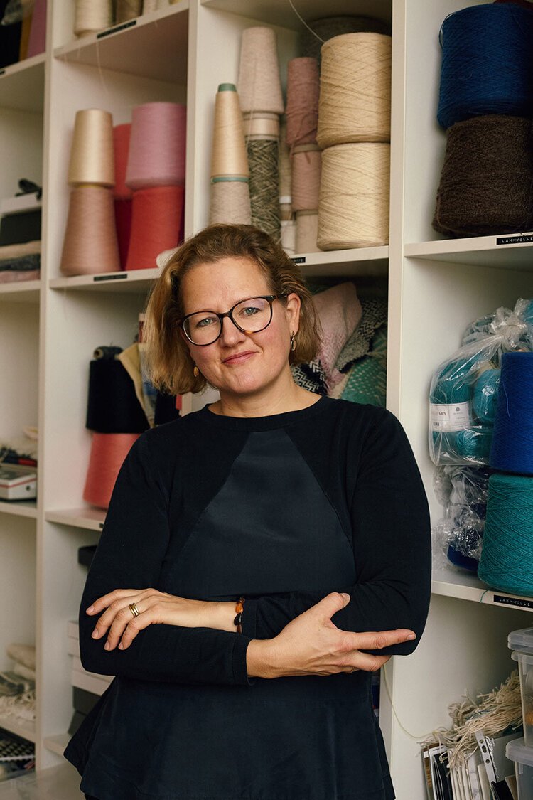 A woman with short light brown hair, glasses, and a black long-sleeve shirt standing in front of shelves filled with colorful spools of yarn and textiles.