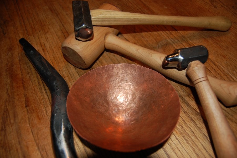 A collection of hammer types, a small copper bowl, and a chisel on a wooden surface.