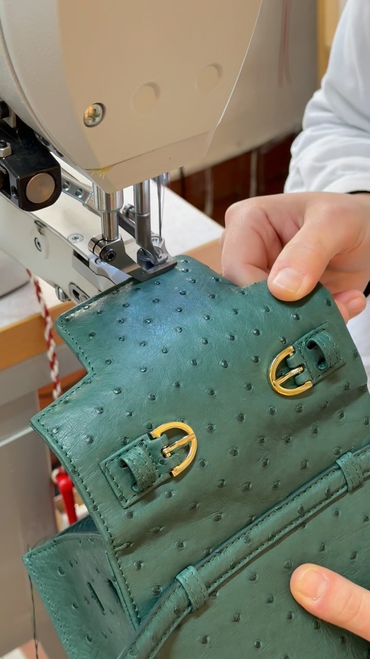 A person sewing a green leather handbag with gold buckles on a sewing machine.