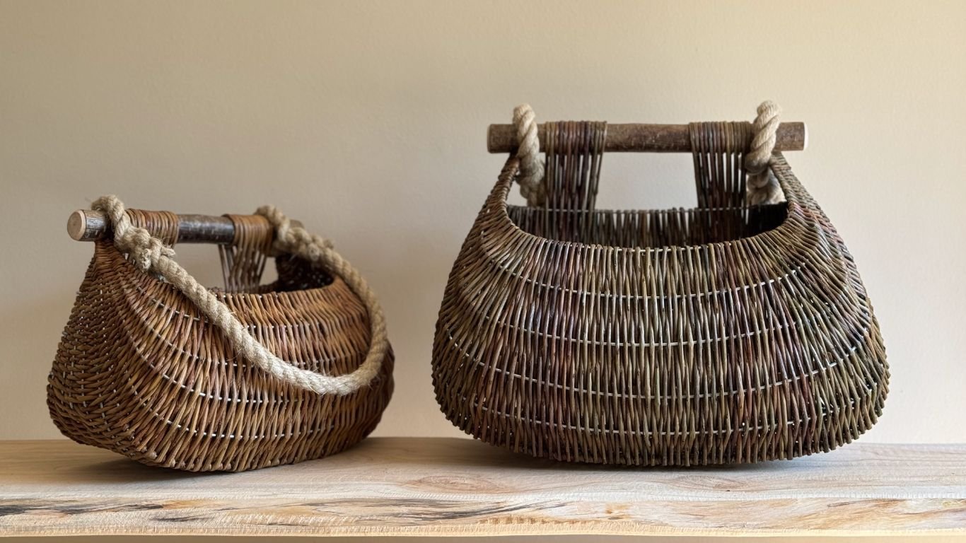 Two handmade woven wicker bags with wooden handles and rope accents, resting on a wooden surface against a beige wall.
