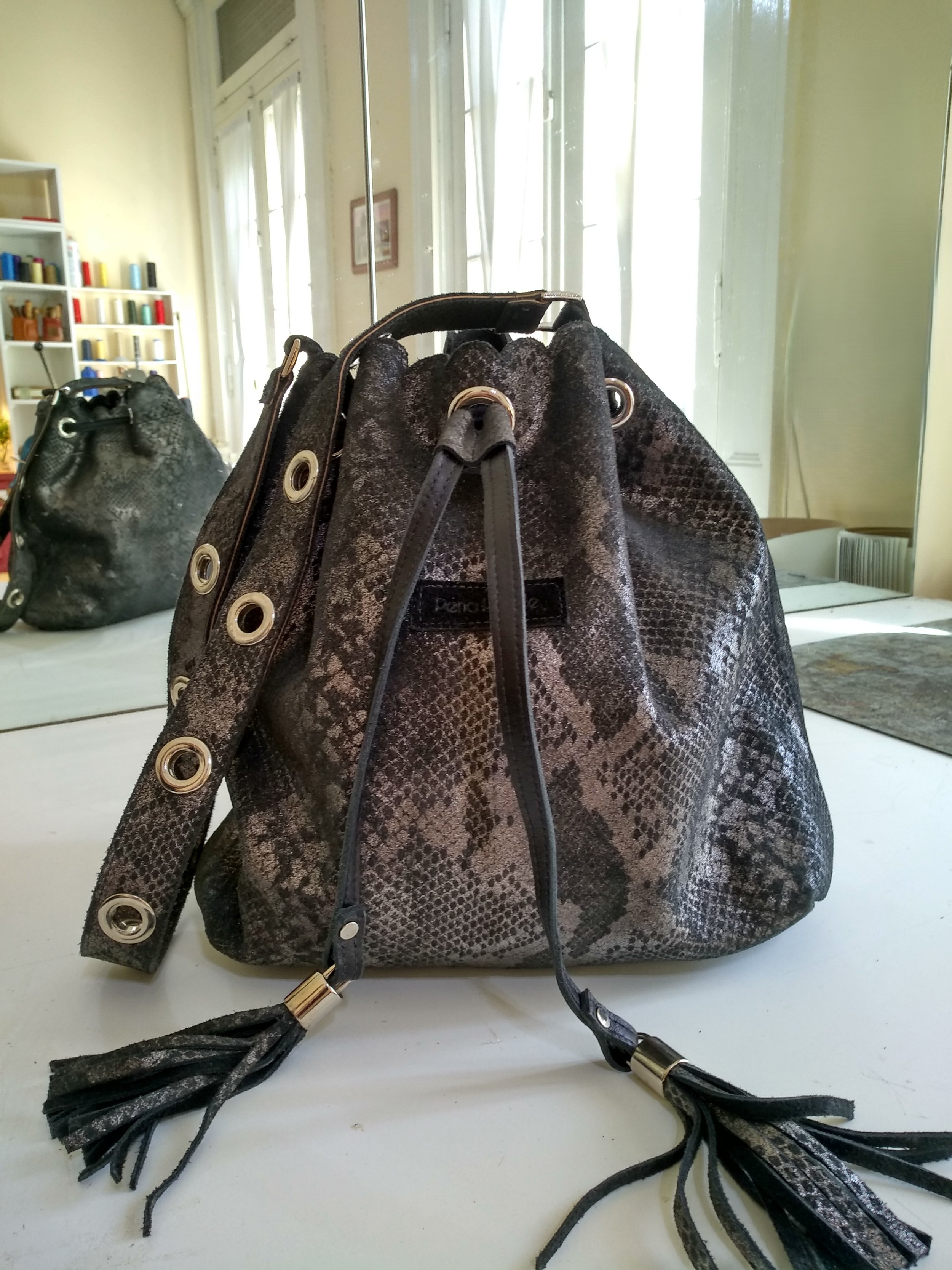 A handmade black and gray snakeskin patterned handbag with drawstring closure and tassel accents, placed on a white surface in front of a mirror, with similar bag visible reflected in the mirror.
