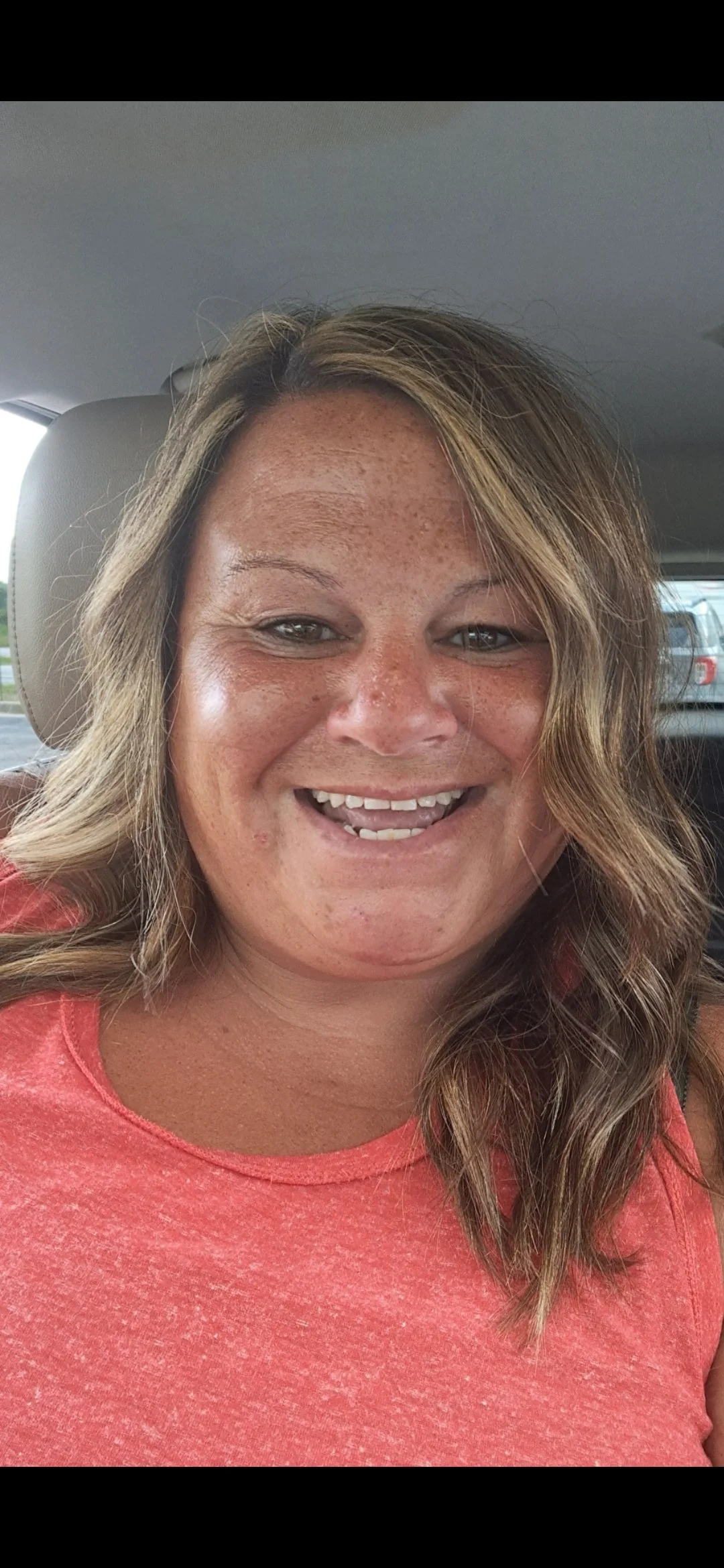 A woman with curly, shoulder-length hair smiling and showing her teeth, wearing a coral-colored top, seated inside a vehicle with a view of cars outside the window.