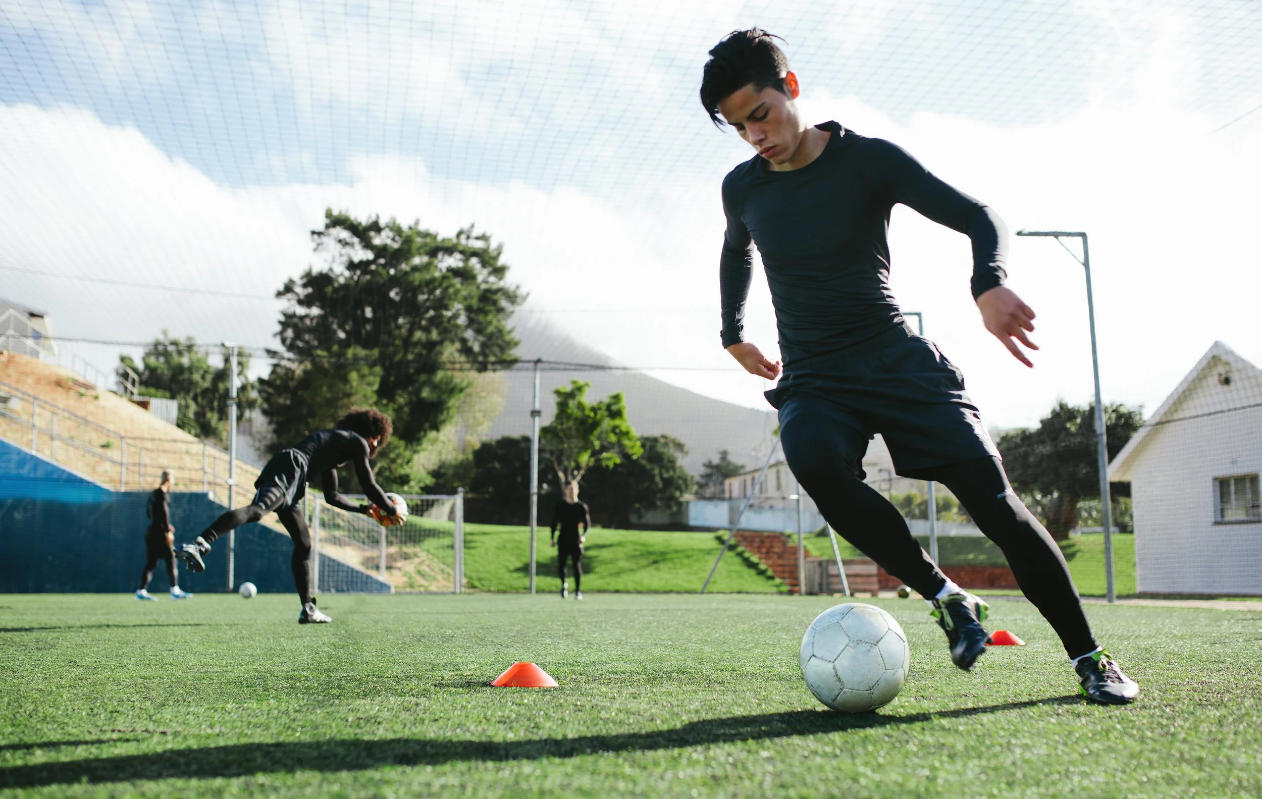 Group of young men practicing soccer drills on a green turf field, with orange cones marking the course, under an overcast sky.