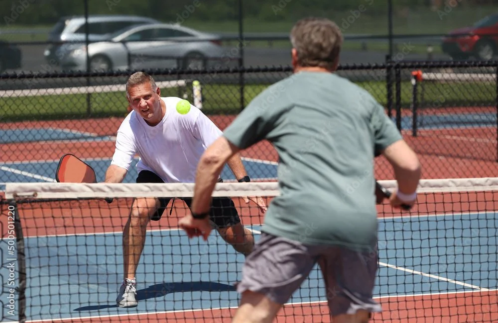 Two men playing pickleball on an outdoor court, one preparing to hit a tennis ball with a paddle while the other stands behind the net.