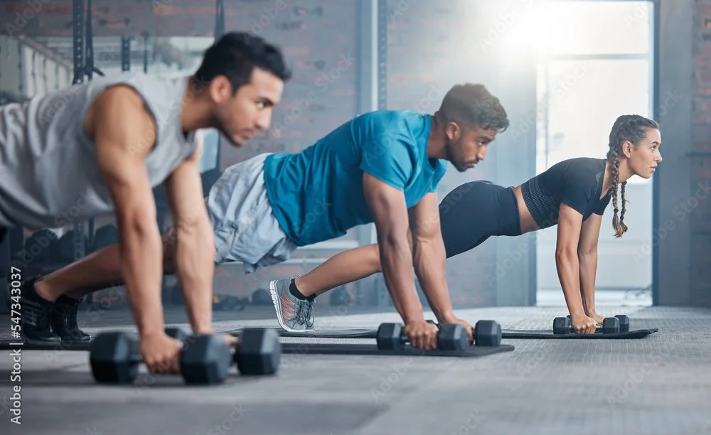Three people doing push-ups with dumbbells in a gym.