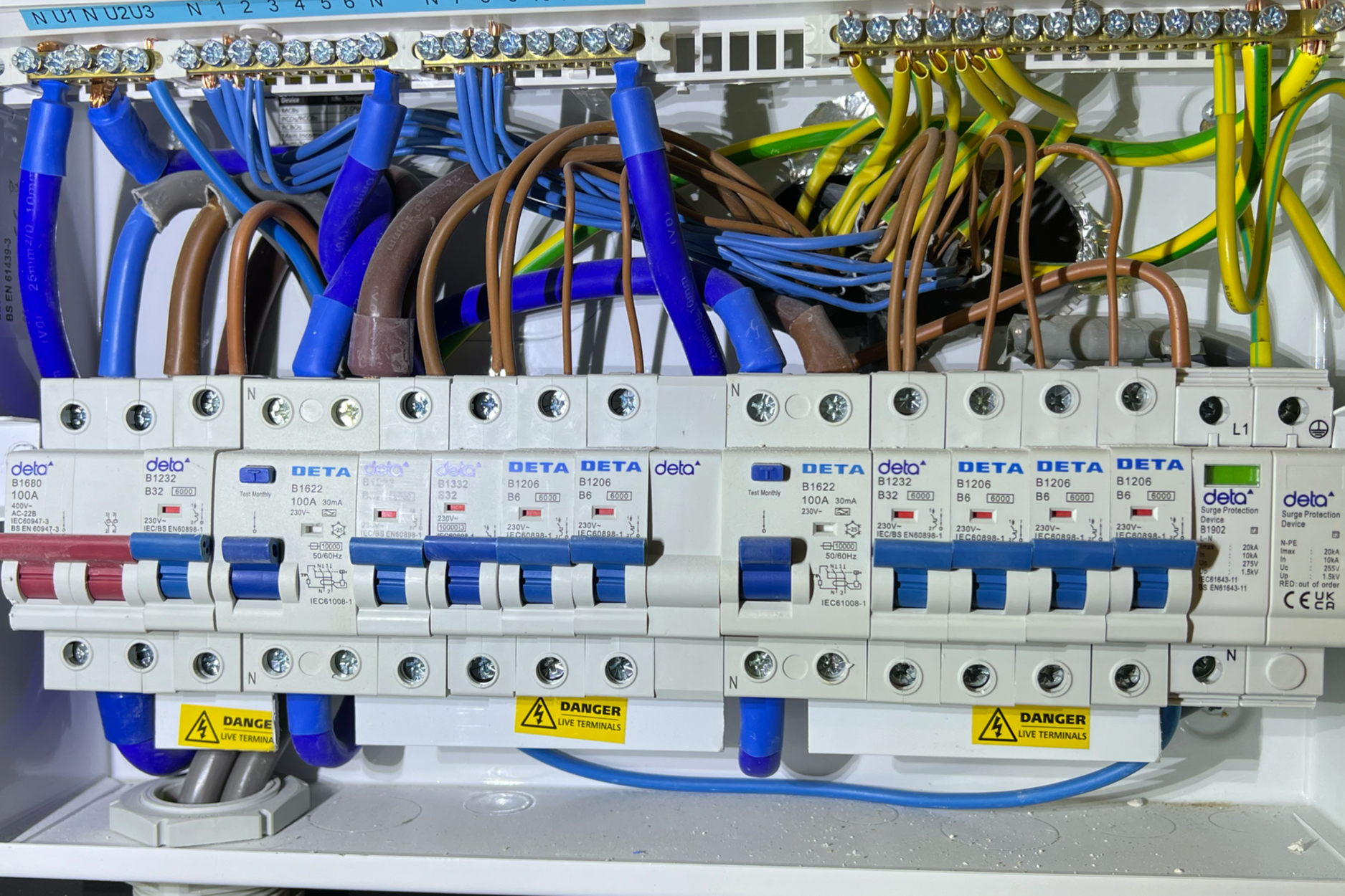 An electrical panel with various blue, brown, and yellow wires connected to circuit breakers. The circuit breakers are labeled with specifications, and there are warning labels indicating live terminals.