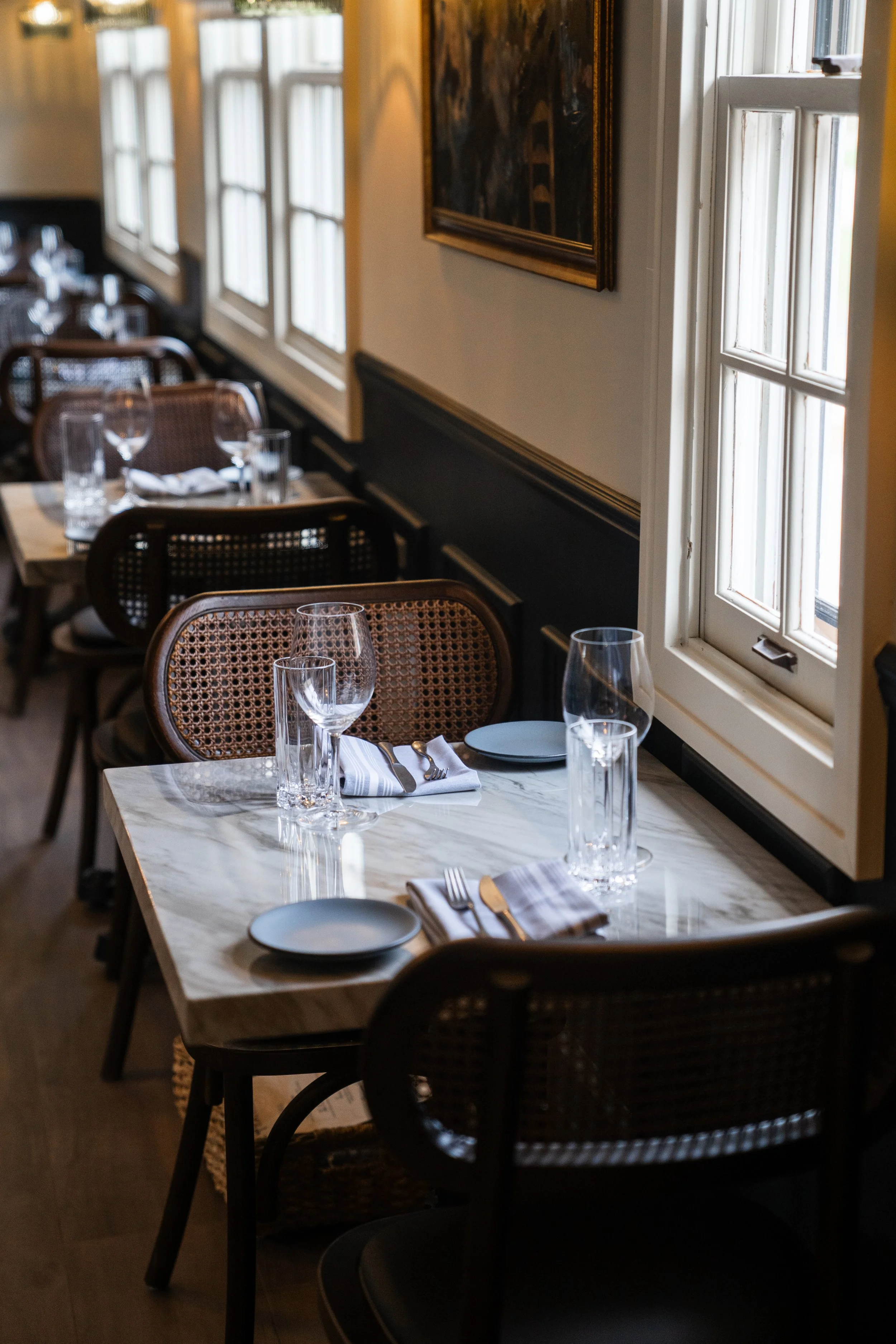 Elegant restaurant interior with marble tables, wicker chairs, and neatly set wine glasses and cutlery near windows.